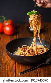 A hand holding a fork with spaghetti with bolognese tomato sauce in black bowl on wooden table
