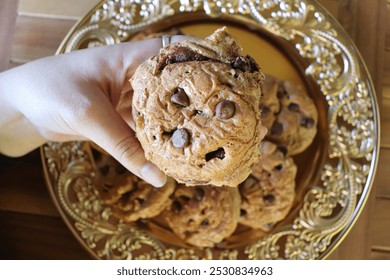 A hand holding a freshly baked chocolate chip cookie over a plate of similar cookies. The cookies are golden brown with chocolate chips, placed on an intricately designed golden rustic plate. - Powered by Shutterstock