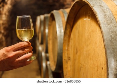 Hand holding a glass of cold white wine in front of oak barrels