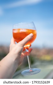 Hand Holding Glass Of Red Aperol Spritz Cocktail Drink With Ice And Slice Of Orange On The Rooftop Bar. Typical Refreshing Drink In Italy. Holiday Concept Photo With Sunset Cityscape In Background.