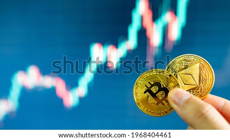 Hand holding gold Bitcoin and Ethereum with blurred candlestick chart in the background. Cryptocurrency and Decentralized finance concept
