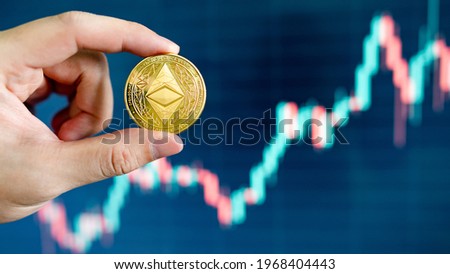 Hand holding gold ETH coin with blurred candlestick chart in the background. Ethereum is a decentralized, open-source blockchain with smart contract. Cryptocurrency and decentralized finance concept