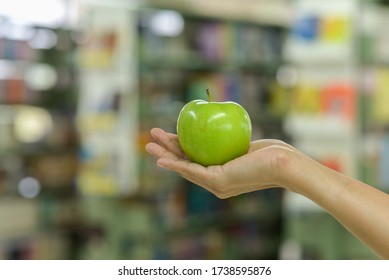 Hand Holding A Green Apple With Blurry Library Background .