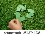 A child’s hand holding a green paper arrow forming a triangular loop on a grassy background. Recycling and environmental conservation, emphasizing youth involvement concept.