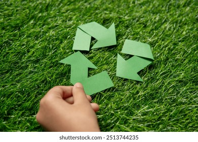 A child’s hand holding a green paper arrow forming a triangular loop on a grassy background. Recycling and environmental conservation, emphasizing youth involvement concept. - Powered by Shutterstock