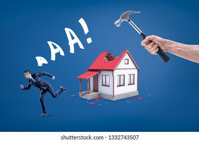 A Hand Holding A Hammer Breaking Small House's Roof With A Businessman Running Away Screaming. Housing Issues. Building Demolition. Lose Home.