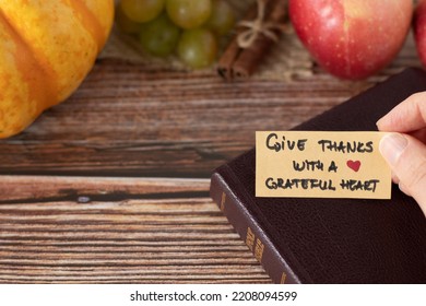 Hand holding a handwritten thanksgiving note to give thanks with a grateful heart with closed Holy Bible Book and fresh autumn fruit on wooden background with copy space. A closeup. Christian concept. - Shutterstock ID 2208094599