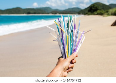 Hand Holding Heap Of Used Plastic Straws On Background Of Clean Beach And Ocean Waves. Plastic Ocean Pollution, Environmental Crisis. Say No Plastic. Single-use Plastic Waste