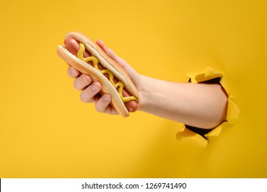 Hand holding a hot dog through a hole in torn yellow paper wall. Delicious street food.