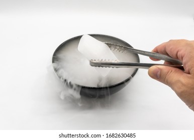 Hand holding ice tweezer with dry ice over black bowl of smoky white in motion isolated on white background.