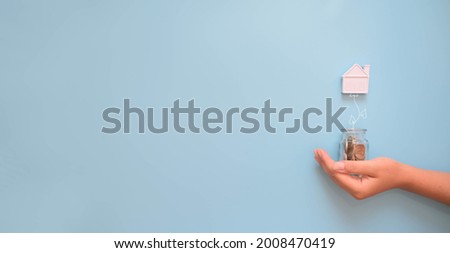 Hand holding a jar with money and miniature house, isolated on blue background. Building, loan, real estate, cost of housing or buying a new home concept.