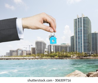 A Hand Is Holding A Key From The New Home. A Concept Of Real Estate Property Agency. Miami Cityscape On The Background.