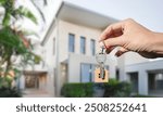 A hand holding a key with a small house-shaped keychain in front of a modern house, symbolizing real estate, home ownership, or moving into a new home.