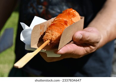 Hand holding Korean style corn dog coated in crispy panko breadcrumbs with hot sauce