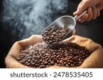 Hand holding a ladle scooping up roasted coffee beans from a pile of beans in burlap sack with cloud of smoke on top. Freshly roasted aromatic strong dark brown coffee beans