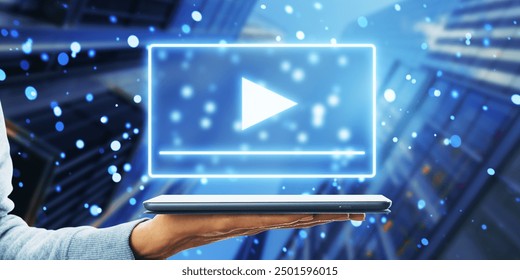 Hand holding a laptop with a streaming play button icon hologram overlay. Cityscape background. Concept of digital media and online content - Powered by Shutterstock