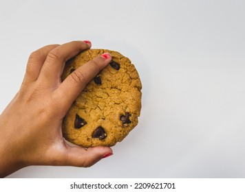 Child’s Hand Holding A Large Chocolate Chip Cookie While Wearing Red Nail Polish Against White Background