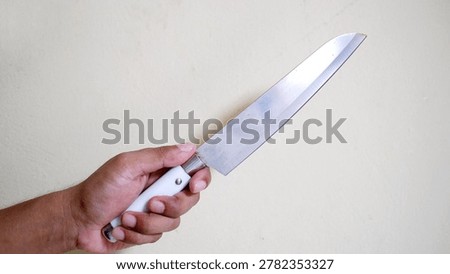 Hand holding a large sharp kitchen knife against a plain wall. Concept of crime, violence, robbery, or domestic threat. Close-up of a weapon held by an aggressor in a dangerous situation.