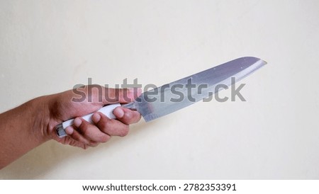 Hand holding a large sharp kitchen knife against a plain wall. Concept of crime, violence, robbery, or domestic threat. Close-up of a weapon held by an aggressor in a dangerous situation.