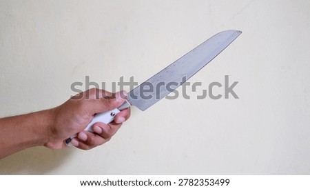 Hand holding a large sharp kitchen knife against a plain wall. Concept of crime, violence, robbery, or domestic threat. Close-up of a weapon held by an aggressor in a dangerous situation.