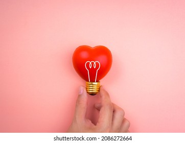 Hand Holding Light Bulb Icon Symbol On Red Heart Ball On Pink Pastel Background, Minimal Style. Love, Care, Sharing, Giving, Wellbeing, Inspiration, And Idea Concepts.