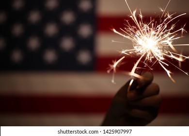 Hand holding lit sparkler in front of the American Flag for 4th of July celebration