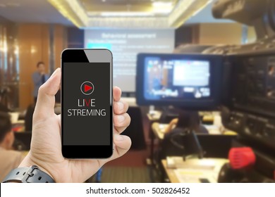 Hand Holding Live Streaming On Smart Phone With Blurred Image Of Video Camera Shooting Business People Meeting In Conference Room