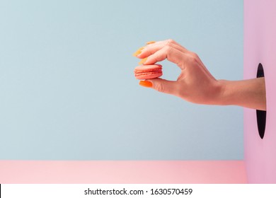 Hand Holding A Macaron Through The Hole In The Wall On Blue Background. Delicious Pastry, Item Of Cafe Menu.