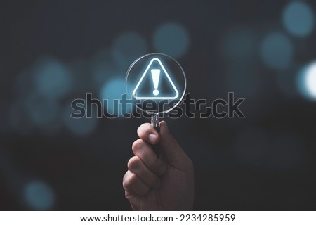 Hand holding magnifier glass with red triangle caution warning sign for notification error and maintenance concept.
