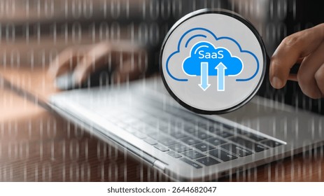 A hand holding a magnifying glass focusing on the SaaS (Software as a Service) cloud icon over a laptop keyboard with a binary code background. Concept of digital cloud computing and analysis.