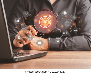 Hand holding a magnifying glass looking at the target with business icons on a virtual screen. Concept of setting a business objective, target audience, reaching the goal and achieving success - Shutterstock ID 2112278027