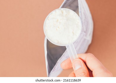 Hand Holding Measuring Spoon With Portion Of Protein Powder Over Open Pack On Brown Table, Top View, Close-up