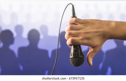 Hand Holding Microphone And Showing Thumbs Down In Front Of A Crowd Of Silhouette People. Failed Public Speaking And Giving Speech Concept. Singing Out Of Tune To Mic, Bad Karaoke Or Poor Talent Show.
