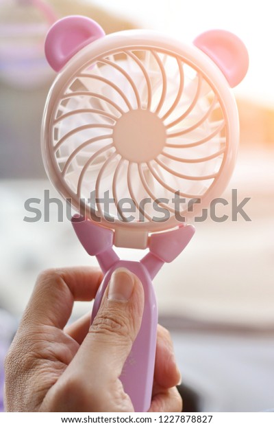 Hand holding mini portable cute electric fan with\
sunlight on blurred background for illustration hot weather or\
summer season.