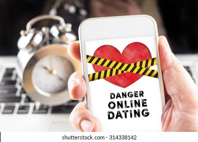 Hand holding mobile with caution tape on heart and Danger online dating  on screen with clock and laptop at background, Internet love concept.