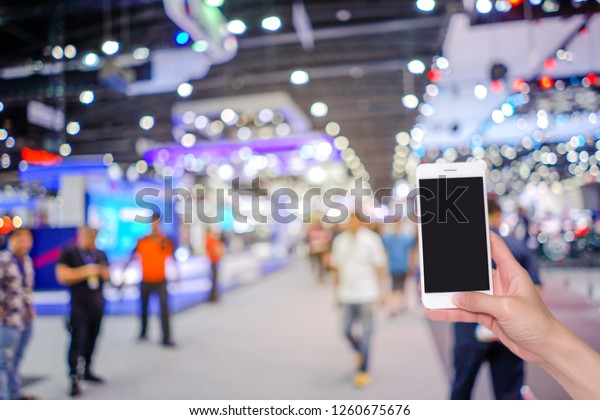 Hand holding mobile phone with\
abstract blurred background image and bokeh light of crowd people\
at cars exhibition show, internet and social network\
concept