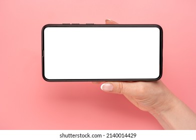 Hand Holding Mobile Phone In Horizontal Position For Mockup. Watching Streaming Video On Cellphone On Pink Table Background. Close Up Of Horizontal Black Smartphone With Blank Screen In Woman Hands