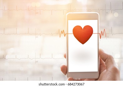 Hand Holding Mobile Phone With Red Heart On Echo Cardiogram Graph Background. In Blur Hospital Room Background.