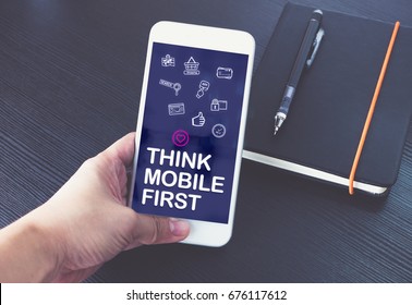 Hand Holding Mobile Phone With Think Mobile First Word And Feature Icon Over Notebook On Black Wood Table Background,Digital Marketing Concept ,office Desk