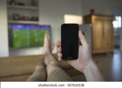 A Hand Holding A Mobile Phone Which Displays A Black Touch Screen. The Background Is A Living Room With A TV Which Displays A Soccer Match. The Person's Feet Are Laying On The Wooden Table.