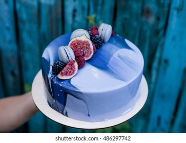 Hand holding ombre marble mousse cake decorated with blue and violet mirror glaze, fruits and macaron. Modern dessert. Wooden hand painted blue background.