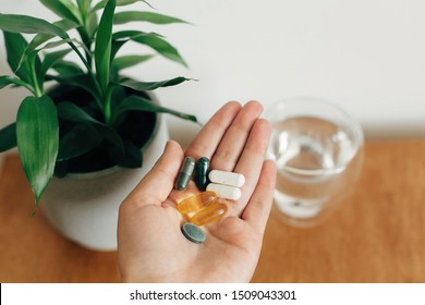 Hand holding omega 3, spirulina, chlorophyll,magnesium  capsules above glass of water on wooden table. Morning vitamin pills. Dietary supplements. Health support. Biologically active additives