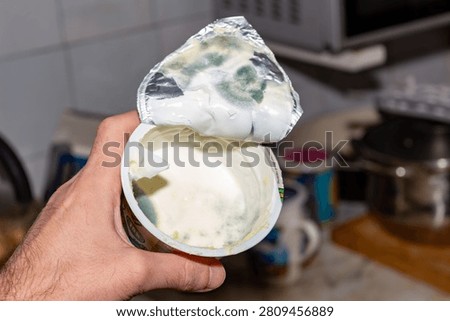 Hand holding open plastic container of spoiled white cream with green mold fungus