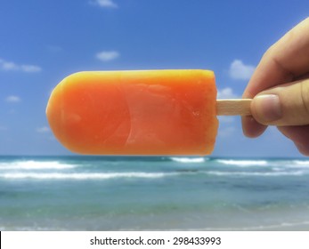 Hand holding an orange popsicle with the sea in the background - Powered by Shutterstock