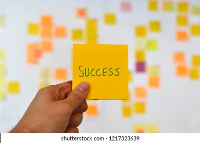 A hand is holding an orange success sticker and there is a Kanban board of agile methodology on the background, which is a developing trend in Information Technology (IT) business.
