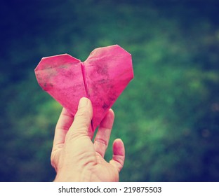  A Hand Holding An Origami Heart Over Green Grass Toned With A Retro Vintage Instagram Filter (shallow Depth Of Field)