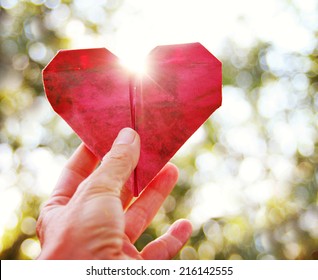 A Hand Holding An Origami Paper Heart Up To The Sun During Sunset