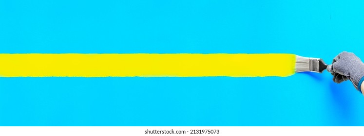 Hand holding paintbrush painting stripe yellow color on blue wall. Colorful art brush strokes painted for home improvement decoration. Horizontal banner backdrop with copy space of Ukraine concept.