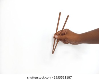 a hand holding a pair of wooden chopsticks against a plain white background. The focus is on the chopsticks, highlighting their smooth texture and natural wooden material. - Powered by Shutterstock