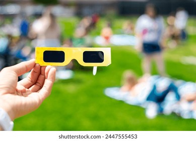 Hand holding paper solar eclipse with blurry crowd people watching totality show picnic yard, Dallas, Texas, April 8, scratch resistant polymer lenses filter out harmful ultraviolet, infrared ray. USA